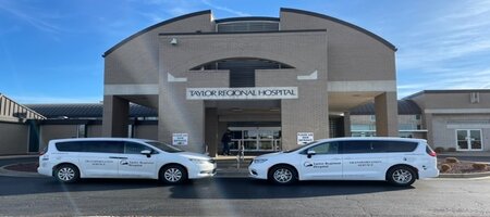 Two Hospitality Van&apos;s parked outside of The Taylor General Hospital facing each other. There is four people smiling (two females) and (two males)